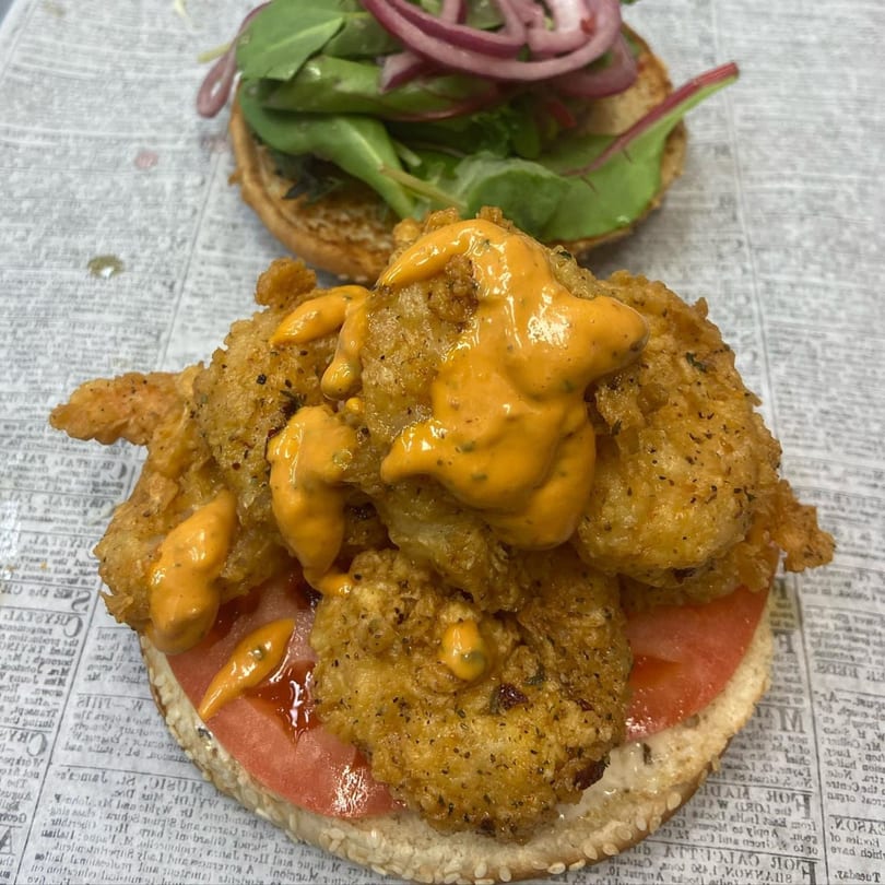 Open-faced fried shrimp sandwich with orange sauce, tomato, and lettuce on a sesame seed bun.