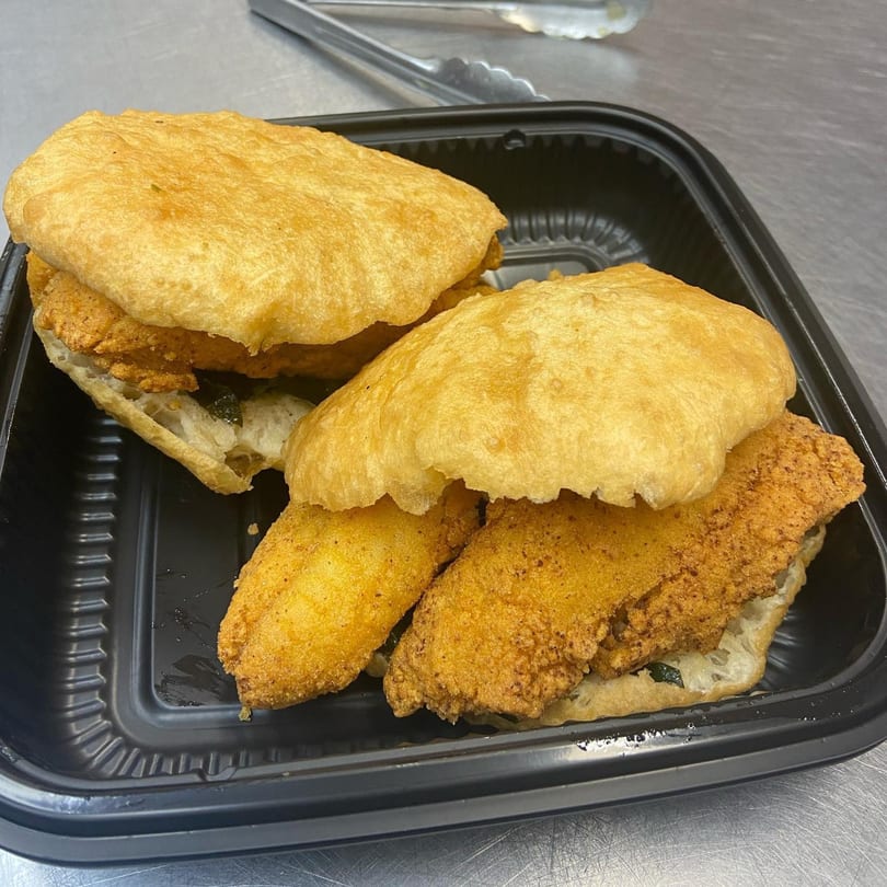 Two golden-fried fish sandwiches on fluffy bread, served in a black takeout container.